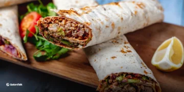 A close up photo of a shawarma filled with thinly sliced chicken, pickles, tomatoes, parsley, and tahini sauce wrapped in pita bread. The shawarma looks freshly made with vibrant ingredients spilling out of the pita. This mouthwatering photo highlights the flavors and textures that make this one of the best shawarmas in Qatar.